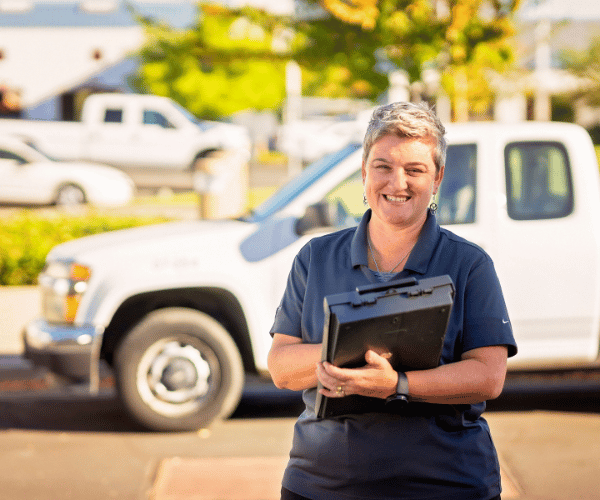 A smiling service worker in a navy uniform stands on a sunny street holding a clipboard.